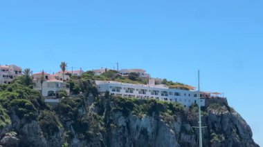 Cala en Porter, Menorca 'daki parlak mavi gökyüzüne bakan yemyeşil ve palmiye ağaçlarıyla çevrili bir tepeye tünemiş beyaz ve pembe evlerin manzarası..