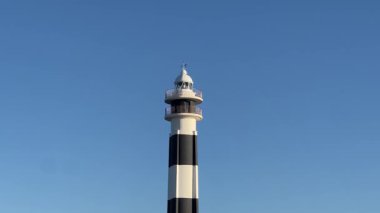 Menorca adasındaki Cap d Artrutx deniz feneri açık mavi gökyüzüne karşı siyah ve beyaz çizgilerle uzun duruyor, Akdeniz kıyısı boyunca gemilere yol gösteriyor..