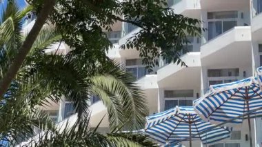 Balkonlu modern beyaz bina, yemyeşil palmiye ağaçları ve mavi gökyüzü ile çevrili, mavi ve beyaz çizgili şemsiyelerle tamamlanmış, sakin bir tropikal atmosfer yaratıyor..