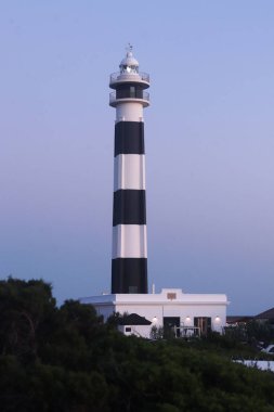 Uzun siyah beyaz çizgili bir deniz feneri beyaz bir binanın yanında, açık mavi bir gökyüzüne karşı duruyor, Cap d Artrutx, Menorca.