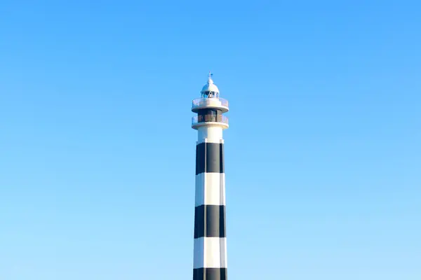 Uzun siyah beyaz çizgili bir deniz feneri beyaz bir binanın yanında, açık mavi bir gökyüzüne karşı duruyor, Cap d Artrutx, Menorca.