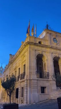 The historic city center of Mahon (Ma), Menorca, features beautiful architecture with elegant buildings, streetlights, and a clear blue sky during the evening.