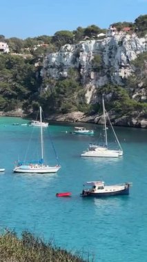 İspanya 'nın Cala Galdana kentinin berrak turkuaz suları ve sakin denizde yavaşça yüzen tekneleriyle nefes kesici manzarası. Çevredeki kayalıklar ve yeşillikler körfezin doğal güzelliğini arttırır..