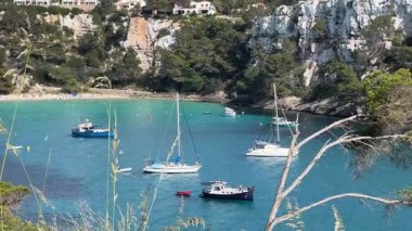 İspanya 'nın Cala Galdana kentinin berrak turkuaz suları ve sakin denizde yavaşça yüzen tekneleriyle nefes kesici manzarası. Çevredeki kayalıklar ve yeşillikler körfezin doğal güzelliğini arttırır..