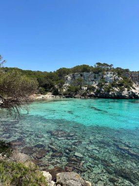 Menorca 'daki Cala Macarella plajının berrak turkuaz suları, uçurumları ve parlak mavi gökyüzünün altında yemyeşil bir manzarası..