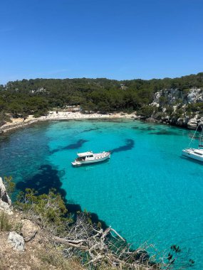 Menorca 'daki Cala Macarella sahilinin kristal berraklığında turkuaz suları. Etrafı çam ağaçları ve kayalık kayalıklarla çevrili. Huzurlu körfezde demirli tekneler var..