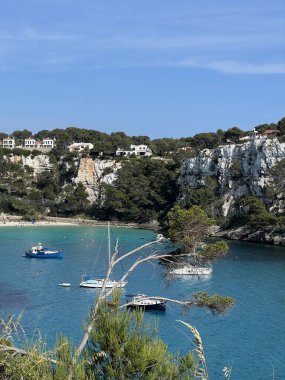 Cala Galdana sahilinin manzarası kristal berraklığında turkuaz su, kayalık kıyılar ve yemyeşil sularla çevrili körfezde huzur içinde yüzen tekneler..