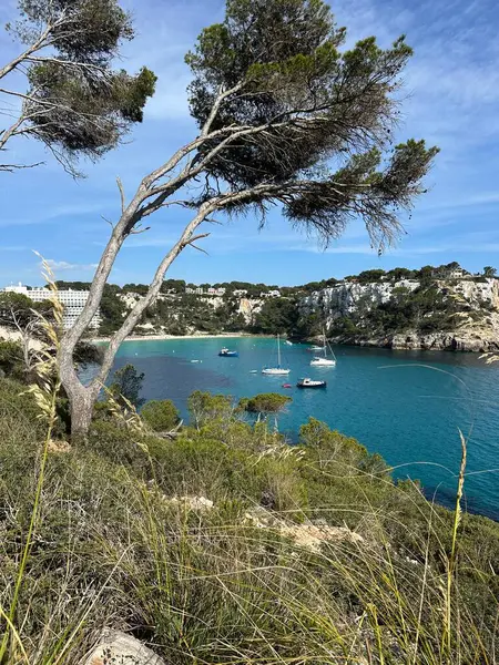 Cala Galdana sahilinin manzarası kristal berraklığında turkuaz su, kayalık kıyılar ve yemyeşil sularla çevrili körfezde huzur içinde yüzen tekneler..