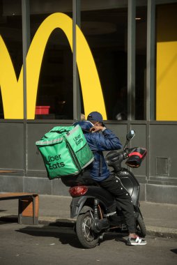 Basel - İsviçre - 7 Mart 2025 - Uber yemek dağıtımı yapan adamın portresi McDonald 's fast food restoranının önünde sokakta scooter üzerinde oturuyor.