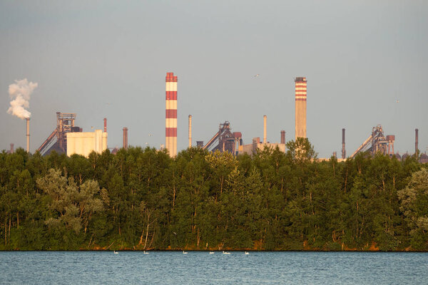 Industrial steel factory, iron works. Metallurgical plant. steelworks. Heavy industry in Europe. Air pollution from chimneys. Ironworks on a background of blue lake.  Nature and industry.