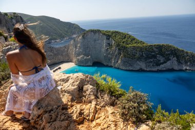 Yunanistan 'da bir uçurumun kenarında oturan beyaz elbiseli güzel kız, Zakynthos adası. Yunanistan 'ın Zakynthos adasındaki Navagio plajı manzarası. Yunanistan 'da Batık Plajı veya Agios Georgios.