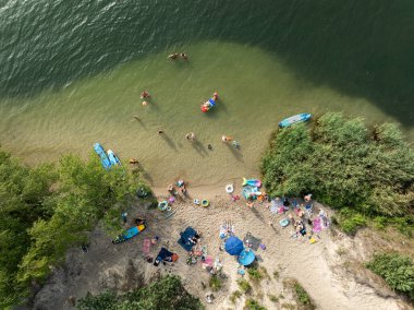 Summer Lake Sahili 'nin hava aracı görüntüsü. Yeşil gölde güneşli bir yaz tatili. Güneşlenmek ve sahilde dinlenmek. Polonya 'daki gölde tatil. İnsanlar göl sahilinde güneşleniyor..