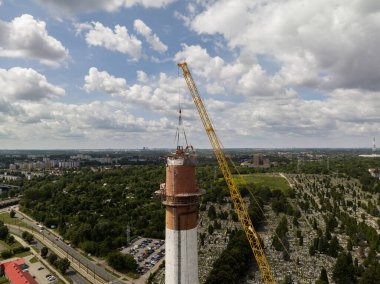 Work on top of the chimney. A suspended excavator on a crane destroys a chimney.Aerial drone view of demolition of chimney, excavator working on smokestack. Small excavator destroying a tall chimney. clipart