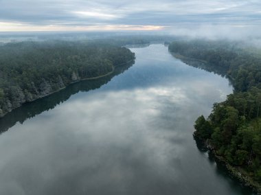 Yeşil ormanla çevrili bir gölün hava aracı görüntüsü. Siste ve bulutlarda büyük bir göl. Kulka Reserve, Masuria, Polonya. Masurian Gölü bölgesinde bir göl. Polonya 'daki Kulka Reserve' de Lesk Gölü.