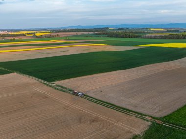 İlkbaharda traktörün ekinleri geniş tarım arazilerine püskürttüğü hava manzarası. Sarı kolza tohumu tarlaları ve yeşil ekinler püskürten bir traktörün insansız hava aracı görüntüsü. Tarım alanları manzarası.