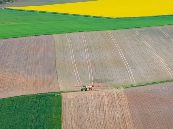 İlkbaharda traktörün ekinleri geniş tarım arazilerine püskürttüğü hava manzarası. Sarı kolza tohumu tarlaları ve yeşil ekinler püskürten bir traktörün insansız hava aracı görüntüsü. Tarım alanları manzarası.