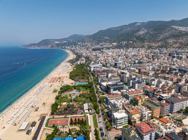 Alanya şehrinin insansız hava aracı görüntüsü, Türkiye. Sahil şeridi, Akdeniz, liman, tepenin üzerindeki tarihi şato ve açık bir yaz gününde arka planda dağların olduğu geniş kentsel alan. Alanya, Türkiye.