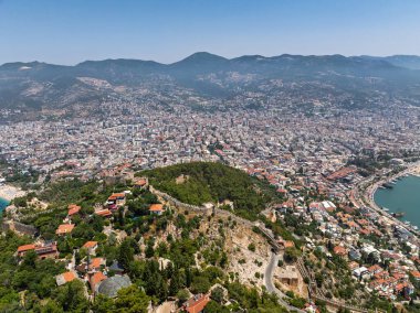 Alanya şehrinin insansız hava aracı görüntüsü, Türkiye. Sahil şeridi, Akdeniz, liman, tepenin üzerindeki tarihi şato ve açık bir yaz gününde arka planda dağların olduğu geniş kentsel alan. Alanya, Türkiye.