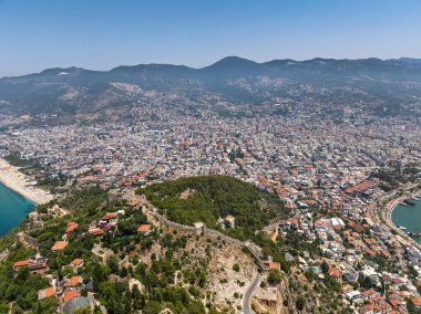 Alanya şehrinin insansız hava aracı görüntüsü, Türkiye. Sahil şeridi, Akdeniz, liman, tepenin üzerindeki tarihi şato ve açık bir yaz gününde arka planda dağların olduğu geniş kentsel alan. Alanya, Türkiye.
