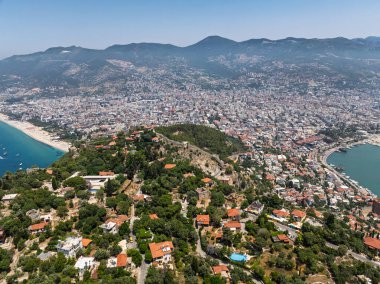 Alanya şehrinin insansız hava aracı görüntüsü, Türkiye. Sahil şeridi, Akdeniz, liman, tepenin üzerindeki tarihi şato ve açık bir yaz gününde arka planda dağların olduğu geniş kentsel alan. Alanya, Türkiye.