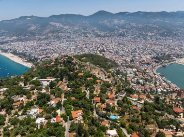 Alanya şehrinin insansız hava aracı görüntüsü, Türkiye. Sahil şeridi, Akdeniz, liman, tepenin üzerindeki tarihi şato ve açık bir yaz gününde arka planda dağların olduğu geniş kentsel alan. Alanya, Türkiye.