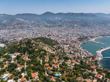 Alanya şehrinin insansız hava aracı görüntüsü, Türkiye. Sahil şeridi, Akdeniz, liman, tepenin üzerindeki tarihi şato ve açık bir yaz gününde arka planda dağların olduğu geniş kentsel alan. Alanya, Türkiye.