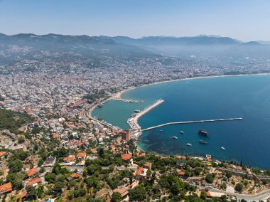 Alanya şehrinin insansız hava aracı görüntüsü, Türkiye. Sahil şeridi, Akdeniz, liman, tepenin üzerindeki tarihi şato ve açık bir yaz gününde arka planda dağların olduğu geniş kentsel alan. Alanya, Türkiye.