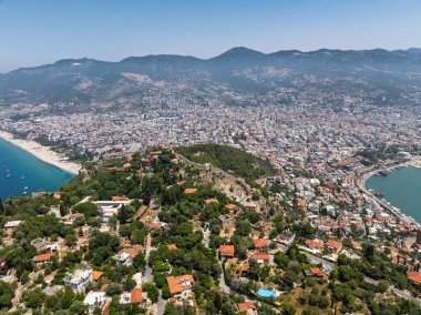 Alanya şehrinin insansız hava aracı görüntüsü, Türkiye. Sahil şeridi, Akdeniz, liman, tepenin üzerindeki tarihi şato ve açık bir yaz gününde arka planda dağların olduğu geniş kentsel alan. Alanya, Türkiye.