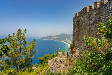 Alanya şehrinin insansız hava aracı görüntüsü, Türkiye. Sahil şeridi, Akdeniz, liman, tepenin üzerindeki tarihi şato ve açık bir yaz gününde arka planda dağların olduğu geniş kentsel alan. Alanya, Türkiye.
