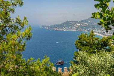 Alanya şehrinin insansız hava aracı görüntüsü, Türkiye. Sahil şeridi, Akdeniz, liman, tepenin üzerindeki tarihi şato ve açık bir yaz gününde arka planda dağların olduğu geniş kentsel alan. Alanya, Türkiye.