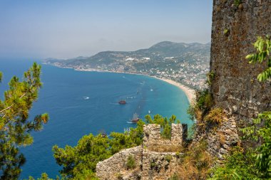 Alanya şehrinin insansız hava aracı görüntüsü, Türkiye. Sahil şeridi, Akdeniz, liman, tepenin üzerindeki tarihi şato ve açık bir yaz gününde arka planda dağların olduğu geniş kentsel alan. Alanya, Türkiye.