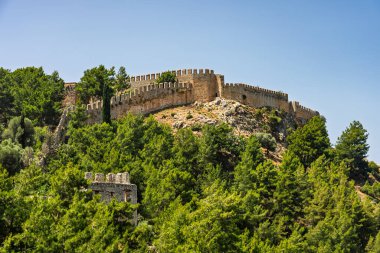 Alanya şehrinin insansız hava aracı görüntüsü, Türkiye. Sahil şeridi, Akdeniz, liman, tepenin üzerindeki tarihi şato ve açık bir yaz gününde arka planda dağların olduğu geniş kentsel alan. Alanya, Türkiye.