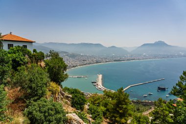 Alanya şehrinin insansız hava aracı görüntüsü, Türkiye. Sahil şeridi, Akdeniz, liman, tepenin üzerindeki tarihi şato ve açık bir yaz gününde arka planda dağların olduğu geniş kentsel alan. Alanya, Türkiye.