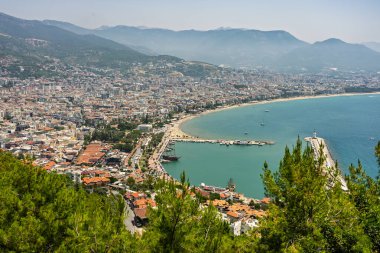 Alanya şehrinin insansız hava aracı görüntüsü, Türkiye. Sahil şeridi, Akdeniz, liman, tepenin üzerindeki tarihi şato ve açık bir yaz gününde arka planda dağların olduğu geniş kentsel alan. Alanya, Türkiye.