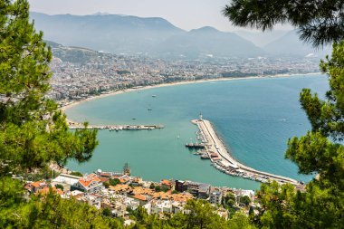 Alanya şehrinin insansız hava aracı görüntüsü, Türkiye. Sahil şeridi, Akdeniz, liman, tepenin üzerindeki tarihi şato ve açık bir yaz gününde arka planda dağların olduğu geniş kentsel alan. Alanya, Türkiye.