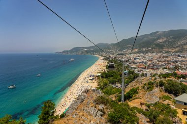 Alanya şehrinin insansız hava aracı görüntüsü, Türkiye. Sahil şeridi, Akdeniz, liman, tepenin üzerindeki tarihi şato ve açık bir yaz gününde arka planda dağların olduğu geniş kentsel alan. Alanya, Türkiye.
