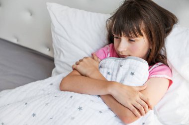 Pijamalı üzgün genç kız yatakta uzanıyor ve kollarıyla kendine sarılıyor. Ateş, yüksek vücut sıcaklığı. Soğuk algınlığı, grip, viral enfeksiyon. Sağlık sigortası konsepti. Hastalık için yatak istirahati. Mevsimsel hastalıklar.