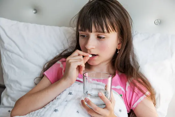 Pijamalı üzgün genç kız yatakta uzanıyor, hap içiyor. Ateş, yüksek vücut sıcaklığı. Soğuk algınlığı, grip, viral enfeksiyon. Sağlık sigortası konsepti. Hastalık için yatak istirahati. Mevsimsel hastalıklar.