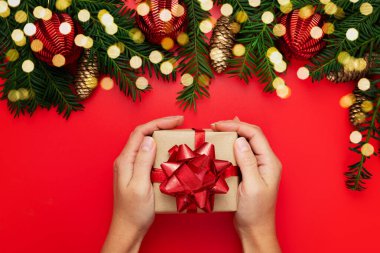 Kraft kağıdına sarılı kırmızı kurdeleli hediye kutusunu tutan kadınların üst görüntüsü. Köknar dalları, koniler, kırmızı toplar. Mutlu Noeller ve yeni yıl. Şenlik havası. 