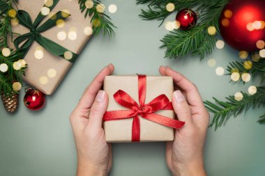 Yeşil arka planda kırmızı kurdeleli hediye kutusunu tutan kadınların üst görüntüsü. Köknar dalları, koniler, kırmızı toplar. Uzayı kopyala Neşeli Noel arkaplanı.