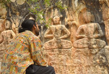 Rear view of a long haired young guy sitting in front of Gyalwa Ringna 5 Dhyani Buddha rock statue which is located in Zanskar, Ladakh, India