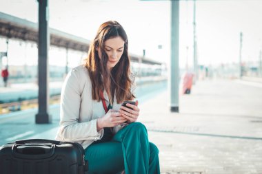 Bir tren istasyonunda beklerken akıllı telefon kullanan gülümseyen iş kadını, tren istasyonunda beklerken cep telefonu kullanan çekici bir genç kız.
