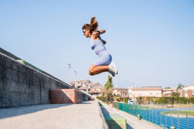 Genç bir kadın stadyum merdivenlerinde hareketli hareketler yapıyor. Spor yaparken güç ve motivasyon saçıyor. Şehir arkaplanı, spor giyim ve güneş gözlükleri aktif yaşam tarzını tamamlıyor.