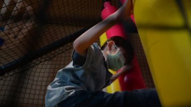 Maskeli bir çocuk otelin çocuk parkından zevk alıyor, kum torbası gibi ağır çekimde boks yapıyor.