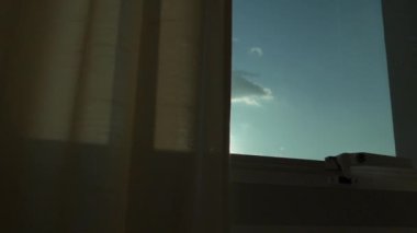Açık pencereden açık gökyüzü görüntüsü kahverengi perdeli, içeriden yavaş çekim sahnesine bakıyor