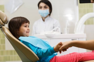 Küçük kız dişçi koltuğunda otururken annesinin elini tutuyor.