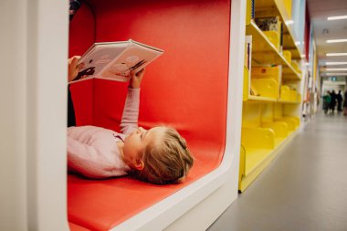 Kütüphanede kitap okurken yerde yatan küçük bir kız. Yüksek kalite fotoğraf. Eğlence konseptini öğreniyorum. Aşk okuma kavramı. 