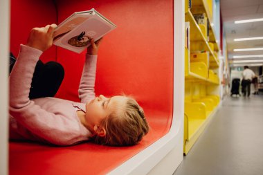 Kütüphanede kitap okurken yerde yatan küçük bir kız. Yüksek kalite fotoğraf. Eğlence konseptini öğreniyorum. Aşk okuma kavramı. 