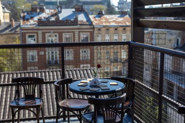 Masası ve sandalyeleri olan rahat bir teras bir Avrupa şehrinin büyüleyici çatılarına ve mimarisine bakıyor. Sabah kahvesi veya sessiz yansıması için huzurlu bir atmosfer yaratıyor..