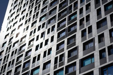 Açık gökyüzünü yansıtan güneş ışığında pencereleri olan yüksek bir apartmanın ön cephesi. Çok katlı yapı, öğlenden sonra aydınlıkta doku, renk ve mimari ritmin bir karışımını gösteriyor..